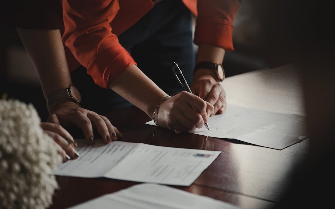 Two people signing IP law documents on a wooden table, with one wearing a red jacket and a watch.