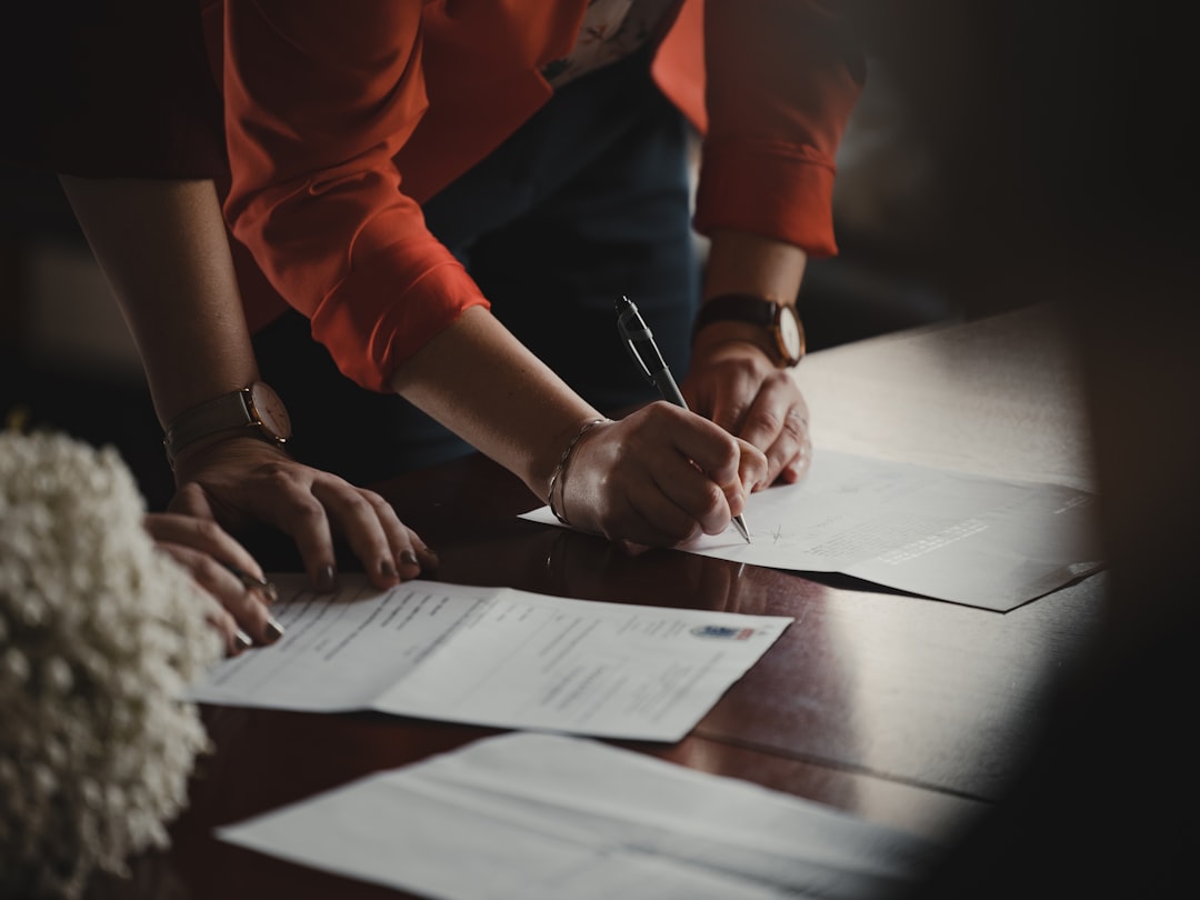 Signing Legal Documents Related to Patent Infringement Two people signing IP law documents on a wooden table, with one wearing a red jacket and a watch.