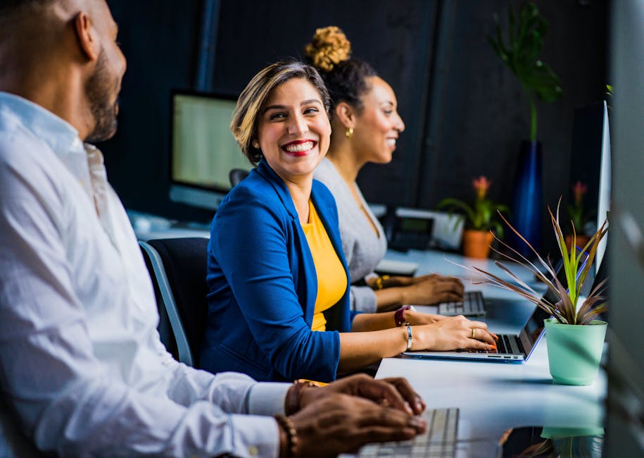 Three people sitting at a desk with laptops, smiling and talking. One woman in a blue blazer is looking at a man.