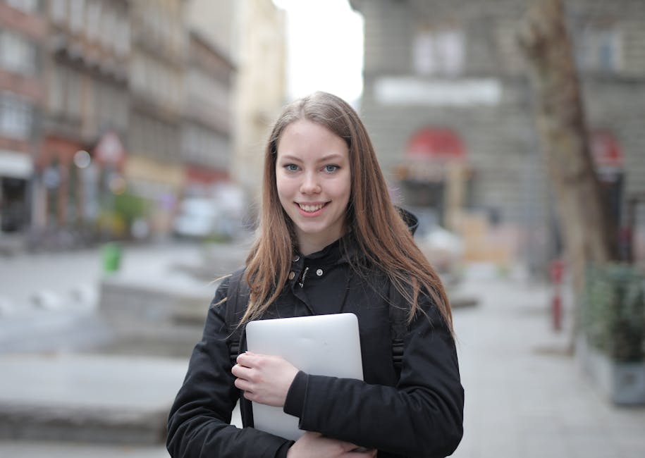 Young woman in a black jacket holding a laptop, standing outdoors in an urban setting.
