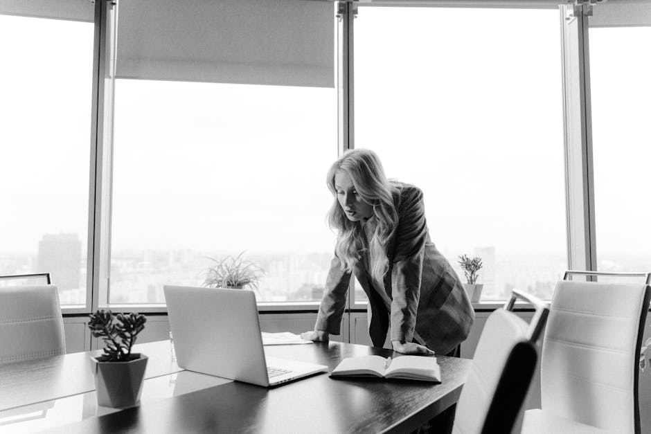Woman in a suit leaning over a table with a laptop and open book in a modern office.