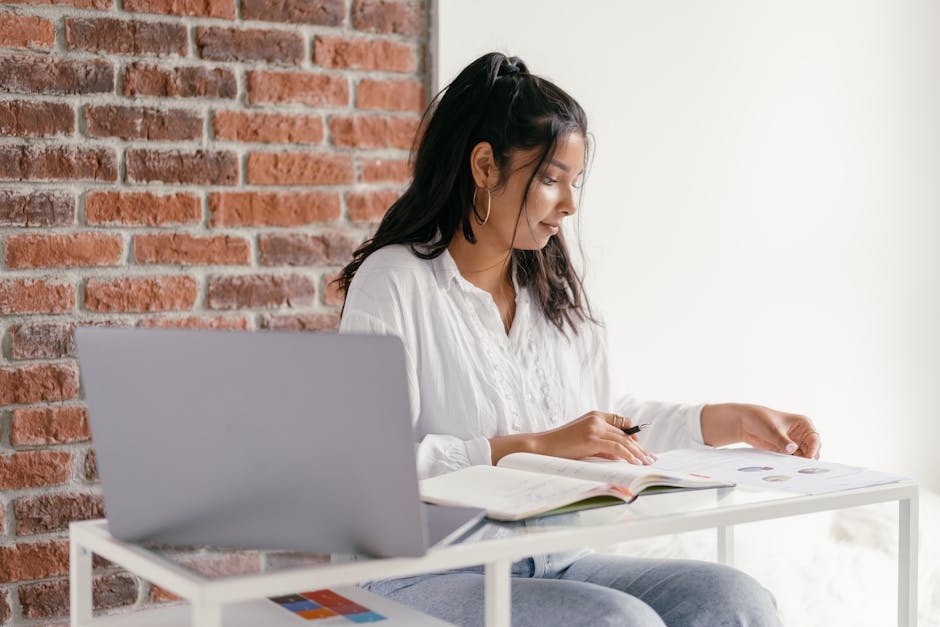 Woman in white blouse studying with books and laptop on a table against a brick wall.