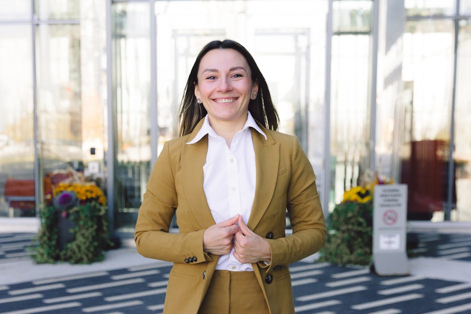 Smiling person in a mustard suit standing outdoors in front of a glass building.