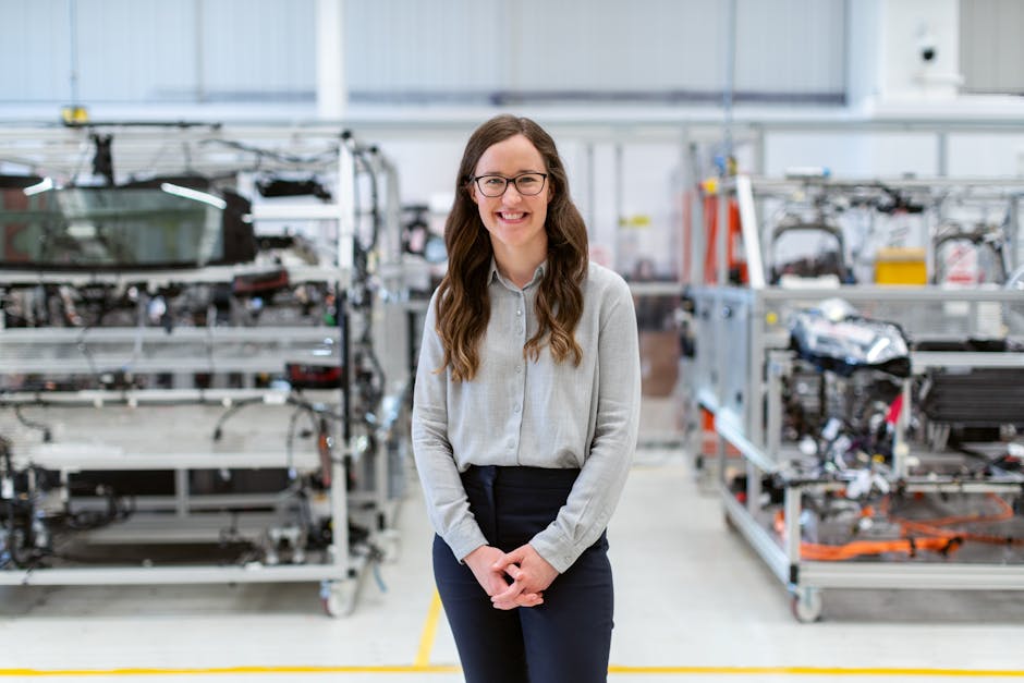 Woman in a grey shirt and glasses standing in a manufacturing facility with machinery in the background.