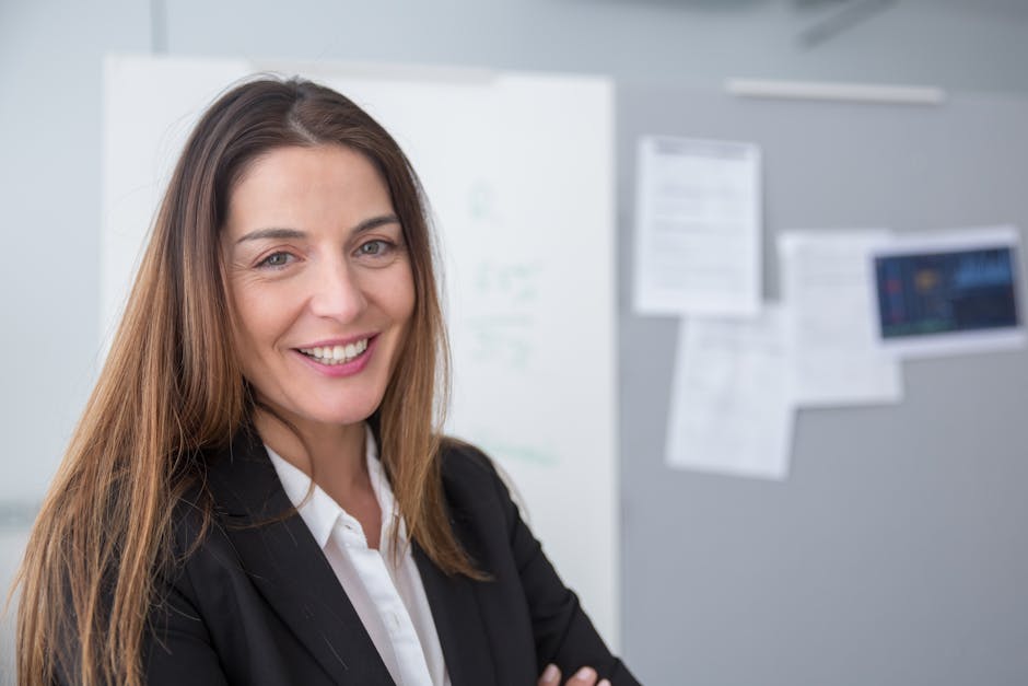 Smiling woman in a black blazer standing in an office with a whiteboard and bulletin board in the background.