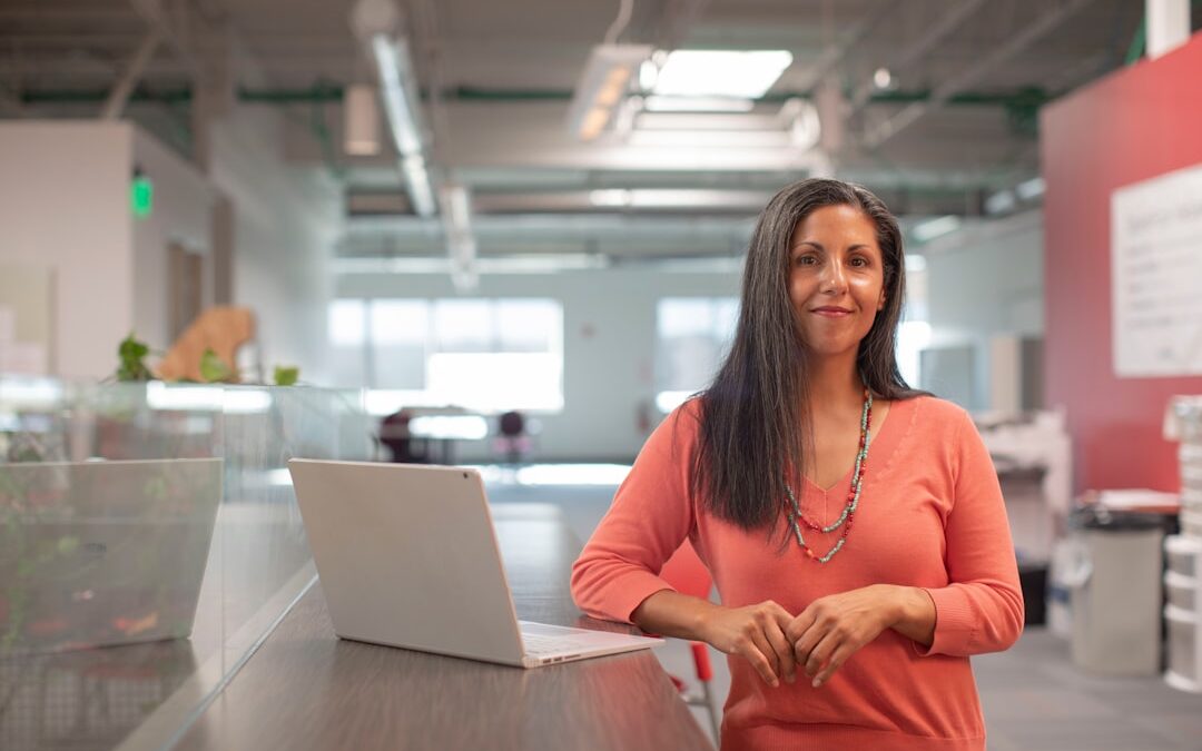 Woman in an orange sweater standing in an office environment with a laptop on a desk.