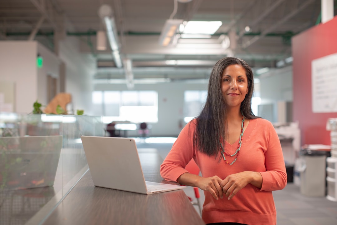 Woman in an orange sweater standing in an office environment with a laptop on a desk.