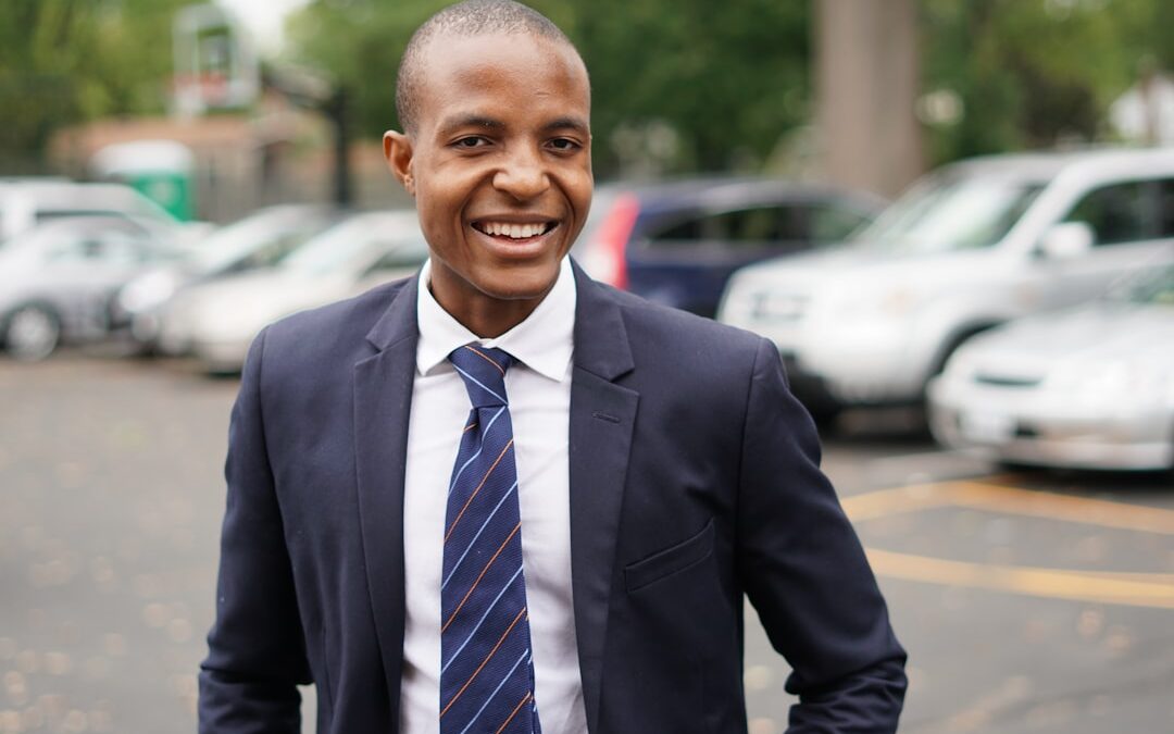 Smiling man in a suit and tie standing in a parking lot with cars and trees in the background.
