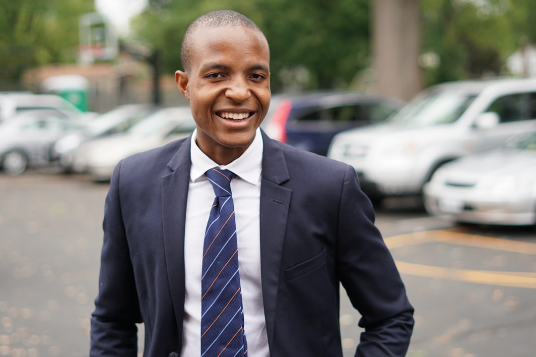 Smiling man in a suit and tie standing in a parking lot with cars and trees in the background.